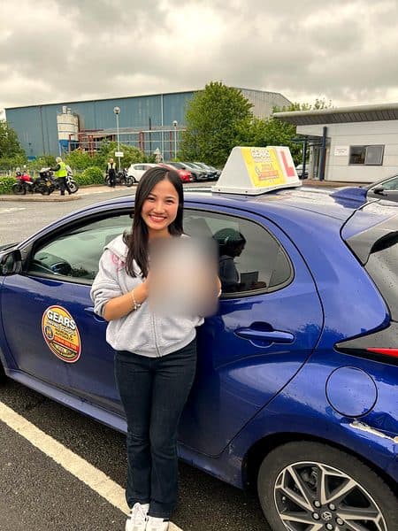 A smiling learner in a grey hoodie holds a pass certificate beside a blue Gears car