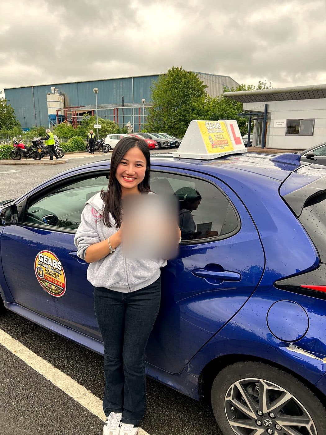 A smiling learner in a grey hoodie holds a pass certificate beside a blue Gears car