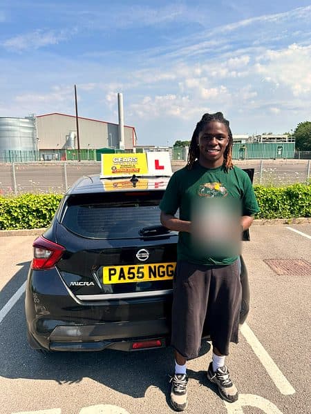A smiling learner in a green T-shirt holds a pass certificate behind a black Nissan Micra