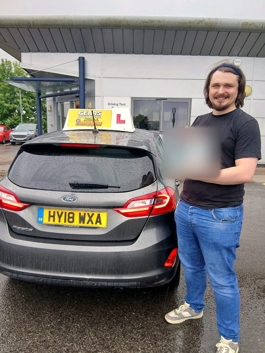 A smiling learner in a black T-shirt holds up a pass certificate beside a grey Ford Fiesta outside the driving test centre