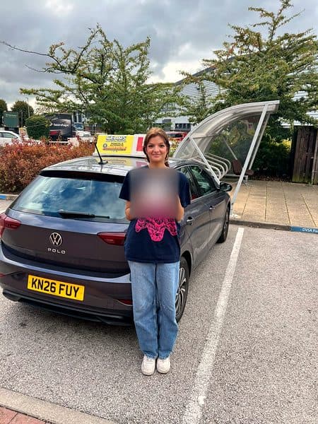 A learner in a dark T-shirt holds a pass certificate behind a grey Volkswagen Polo