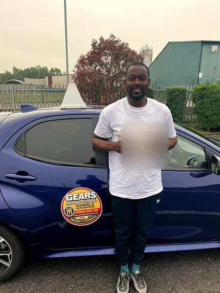 A smiling learner in a white T-shirt holds a pass certificate beside a blue Gears car