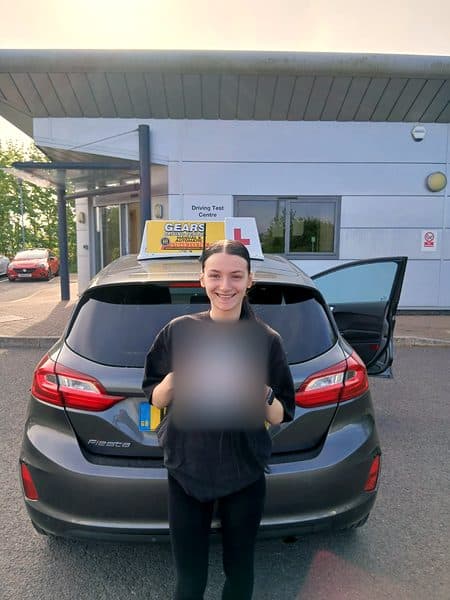 A smiling learner dressed in black holds a pass certificate behind a grey Ford Fiesta outside the driving test centre