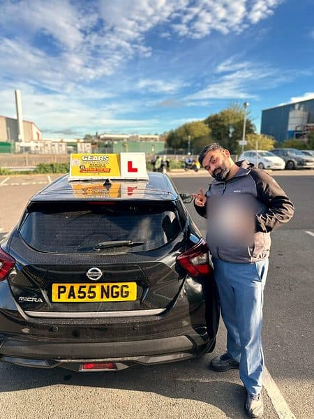 A learner in a grey fleece points at the camera, pass certificate in hand, beside a black Nissan Micra under a blue sky