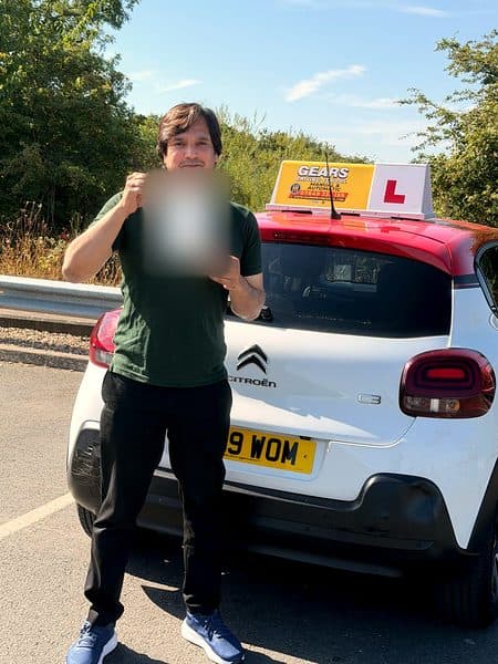 A learner in a green polo shirt holds up a pass certificate behind a white Citroën C3 with a red roof