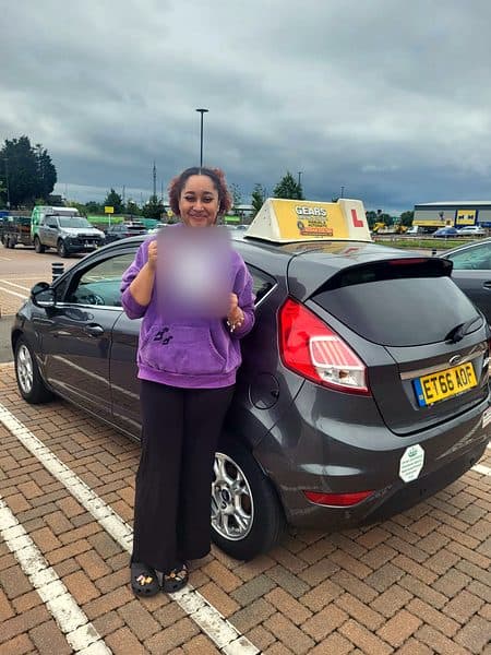 A learner in a purple hoodie holds up a pass certificate beside a grey Ford Fiesta