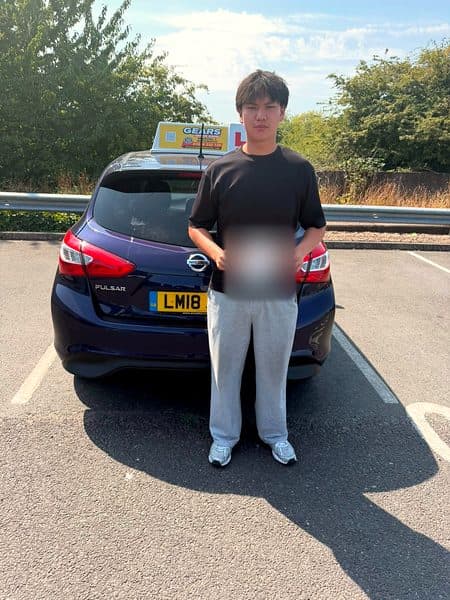 A learner in a black T-shirt holds a pass certificate behind a blue Nissan Pulsar