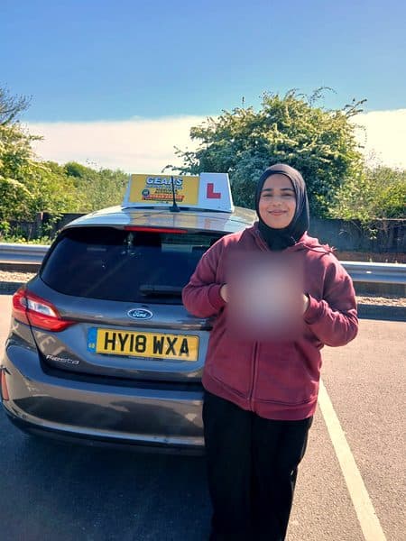 A learner in a hijab and a maroon hoodie holds a pass certificate behind a grey Ford Fiesta