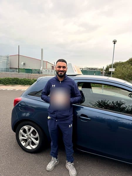 A smiling learner in a navy tracksuit holds a pass certificate beside a blue hatchback
