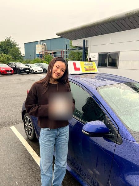 A learner in a brown jumper holds a pass certificate beside a blue Gears car outside the driving test centre