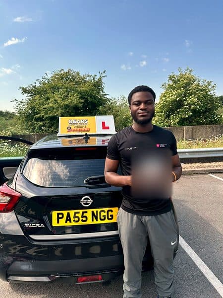 A learner in a black T-shirt holds a pass certificate behind a black Nissan Micra