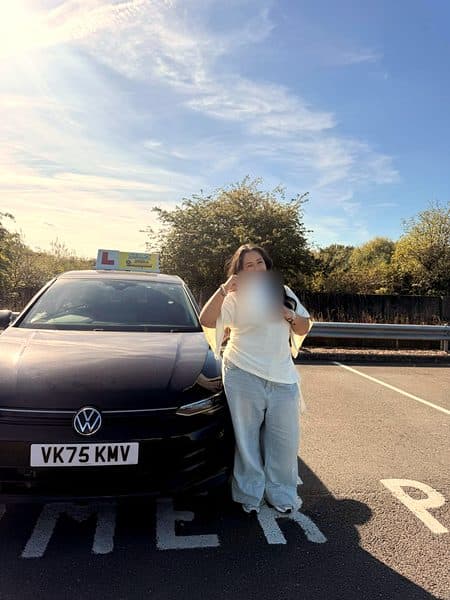 A learner in a cream top holds up a pass certificate beside a black Volkswagen with a Gears roof sign