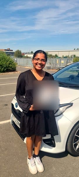 A smiling learner in glasses and a black dress holds a pass certificate beside a white car
