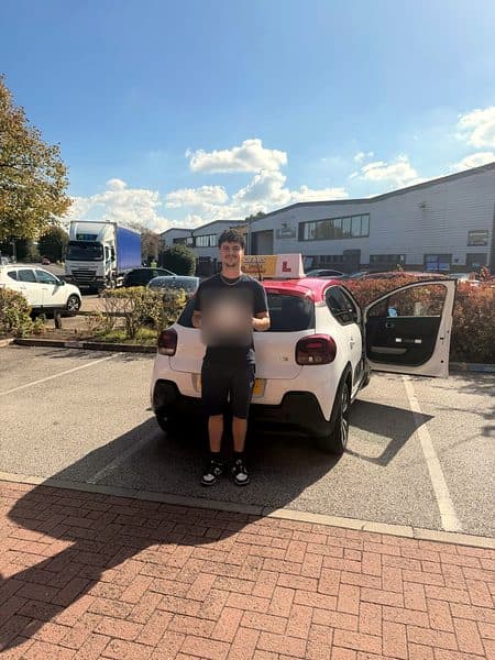 A learner dressed in black holds a pass certificate in front of a white Citroën C3 with a red roof