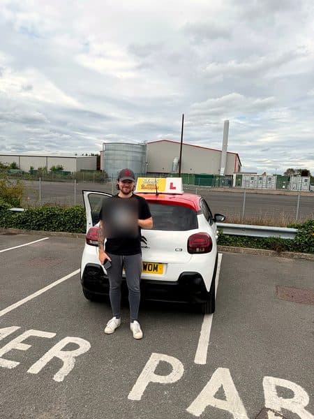 A learner in a cap holds a pass certificate in front of a white Citroën C3