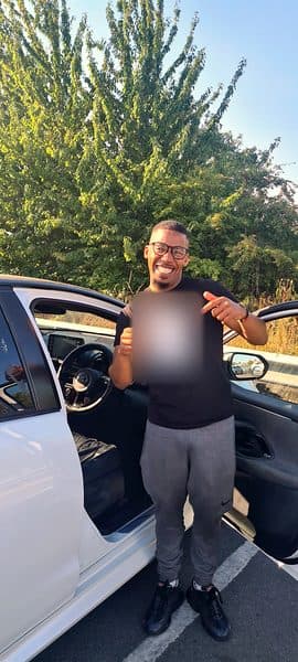 A beaming learner in glasses points at a pass certificate beside the open door of a white car