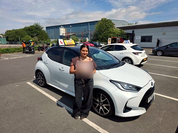 A learner holds a pass certificate in front of a white hatchback in the test centre car park