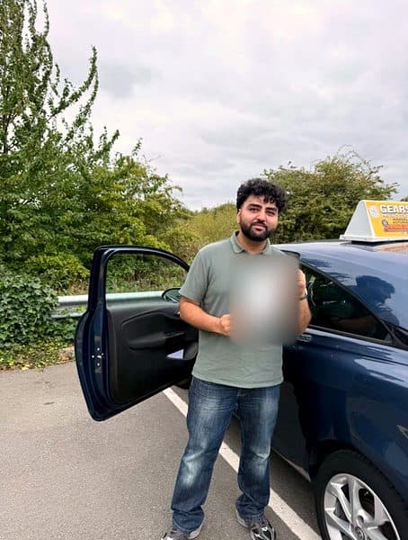 A learner in a green polo shirt holds a pass certificate beside a blue Gears car